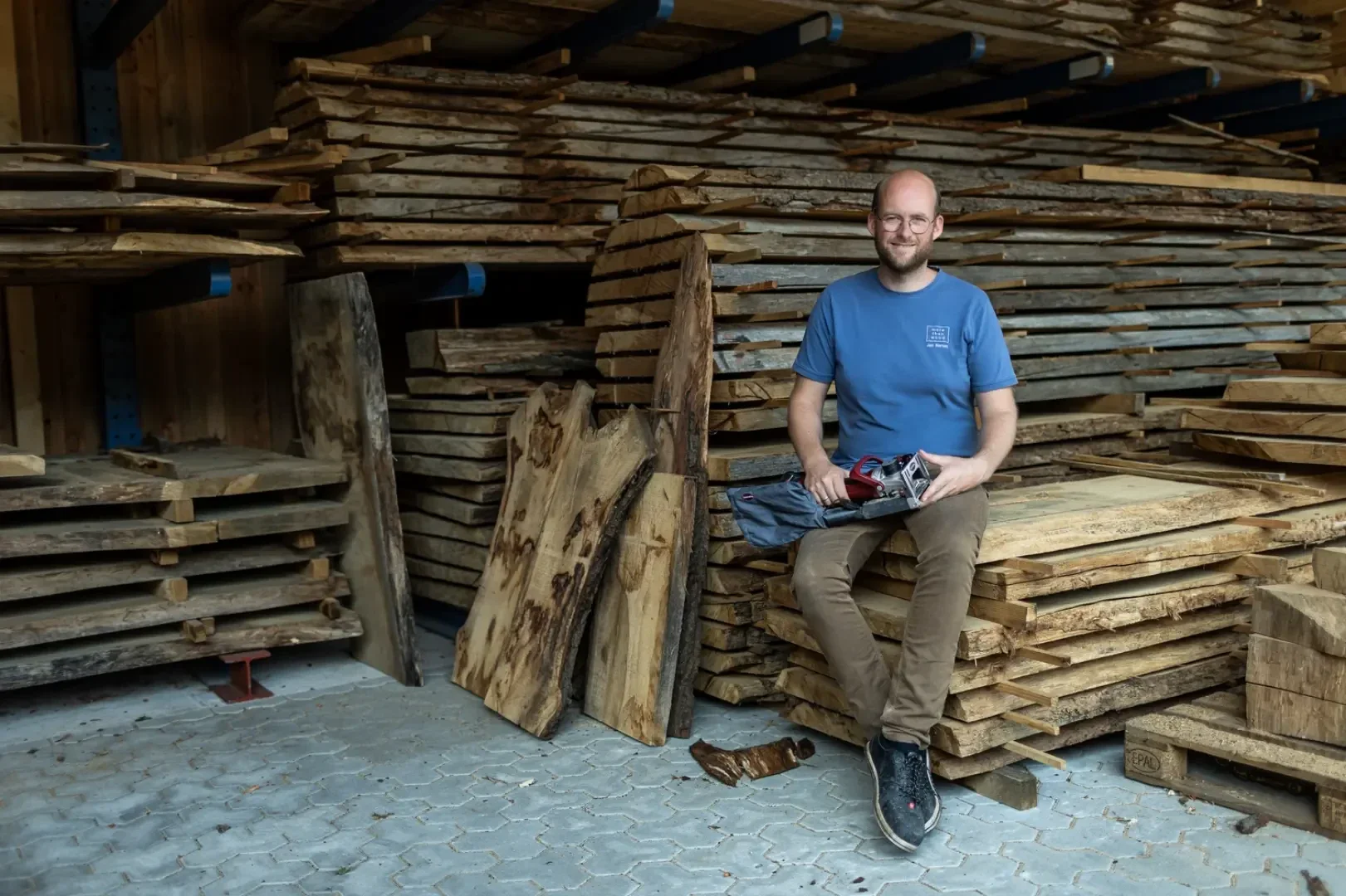 Ein lächelnder Mann in einem blauen Hemd und einer braunen Hose sitzt auf gestapelten Holzbrettern in einem Holzlager und hält ein Elektrowerkzeug in der Hand. Um ihn herum sind weitere rohe Holzbretter zu sehen.