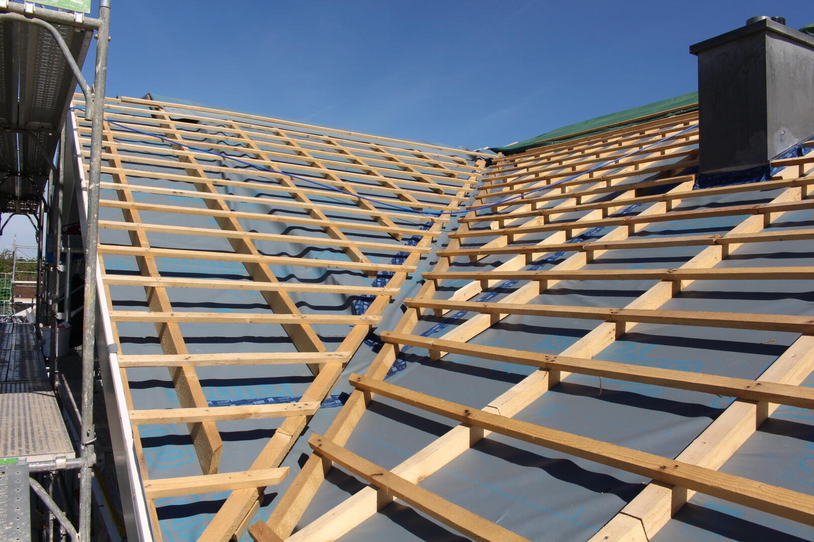 Un toit de maison en pente en cours de construction, recouvert d'une membrane d'étanchéité grise et d'une charpente en lattes de bois, avec un échafaudage à gauche et une cheminée visible à droite, le tout sous un ciel bleu limpide.