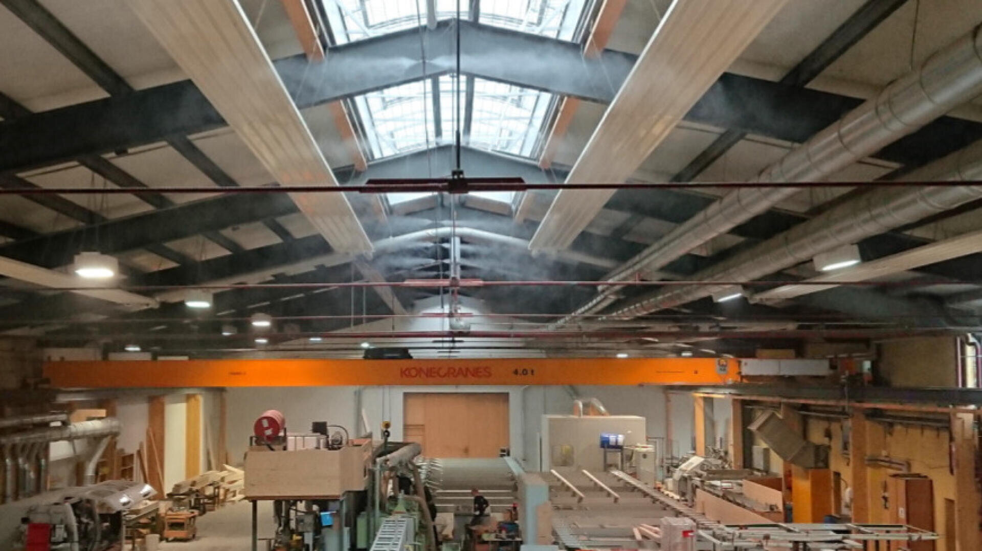 Ein geräumiges Industrielager mit hohen Decken, großen Oberlichtern, freiliegenden Balken und einem orangefarbenen Konecranes-Kran in der Decke. Verschiedene Maschinen, Geräte und Holzmaterialien sind in der Werkstatt verteilt.