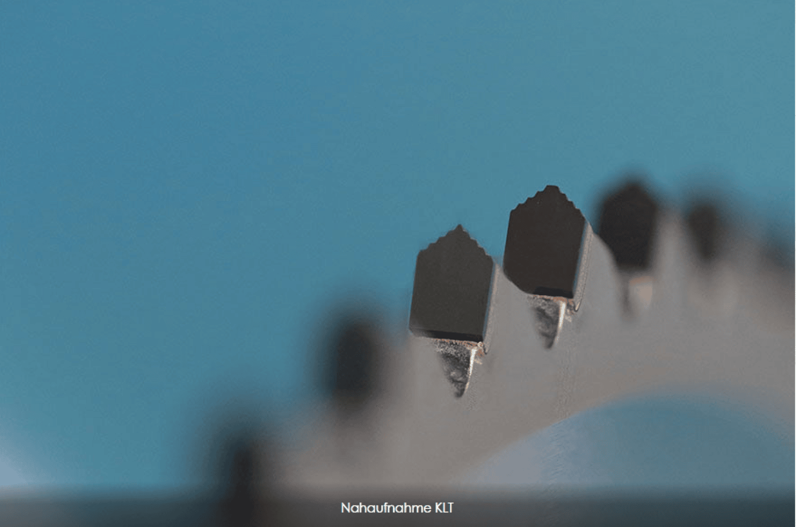 Gros plan de la lame dentelée d'une scie circulaire sur un fond bleu, avec mise au point des dents acérées. Le texte Nahaufnahme KII apparaît en bas.