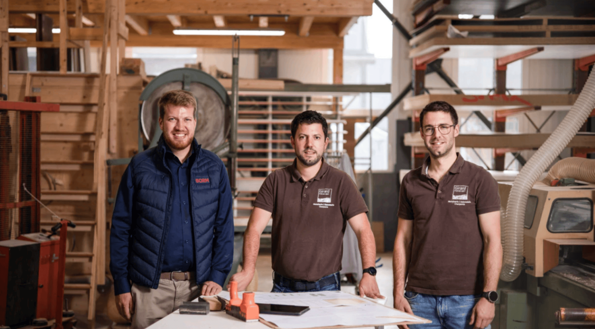 Drei Männer stehen in einer Holzwerkstatt und lächeln in die Kamera. Zwei von ihnen tragen braune Hemden mit Firmenlogos, einer trägt eine marineblaue Weste. Sie stehen hinter einem Tisch mit Papieren und Werkzeugen. Im Hintergrund sind Regale und Geräte zu sehen.