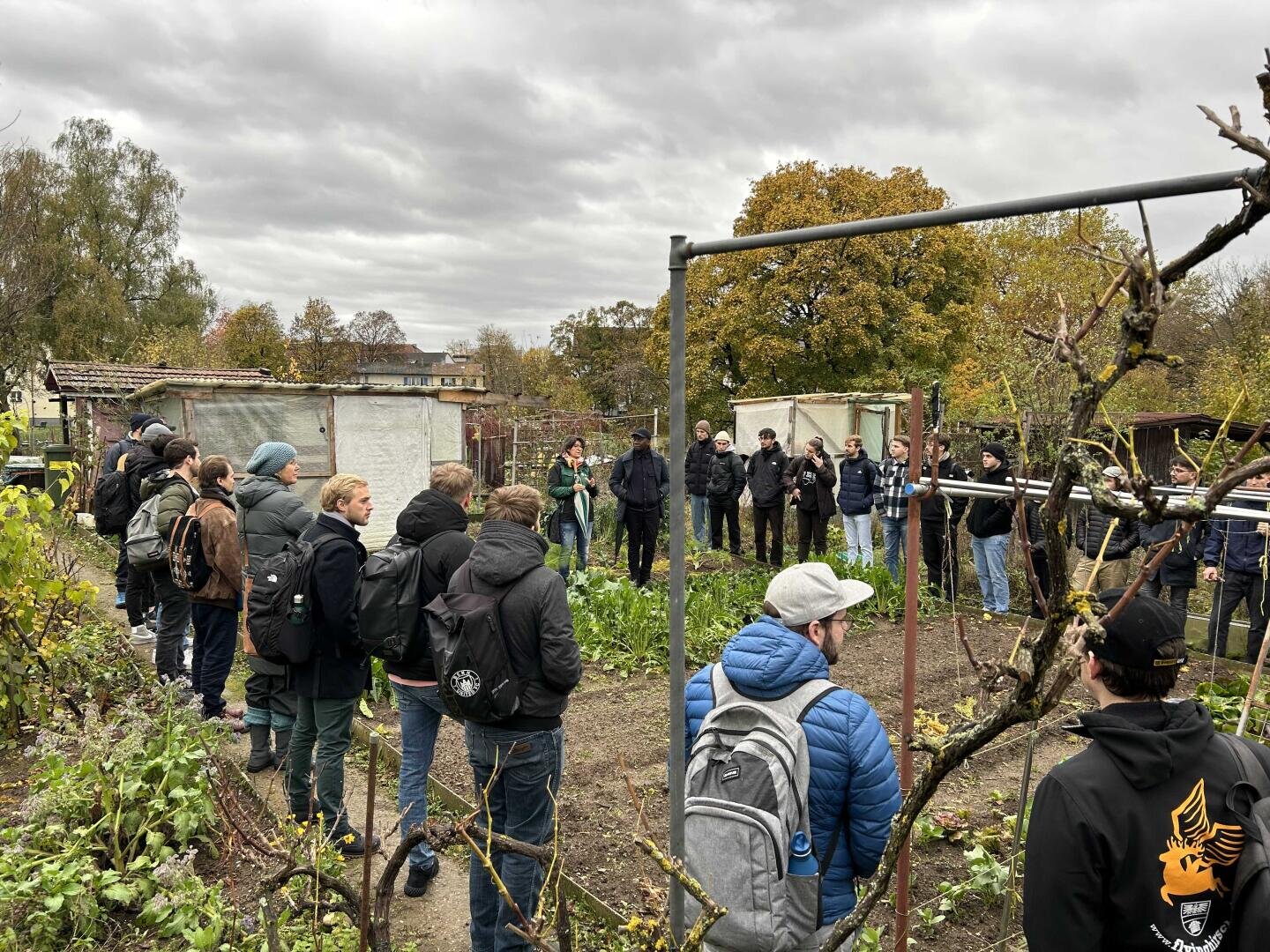 Eine Gruppe von Menschen hat sich an einem bewölkten Tag in einem Gemeinschaftsgarten versammelt und hört einem Redner zu. Sie sind warm gekleidet, umgeben von Pflanzen, Bäumen mit Herbstlaub und kleinen Gartenhäuschen.