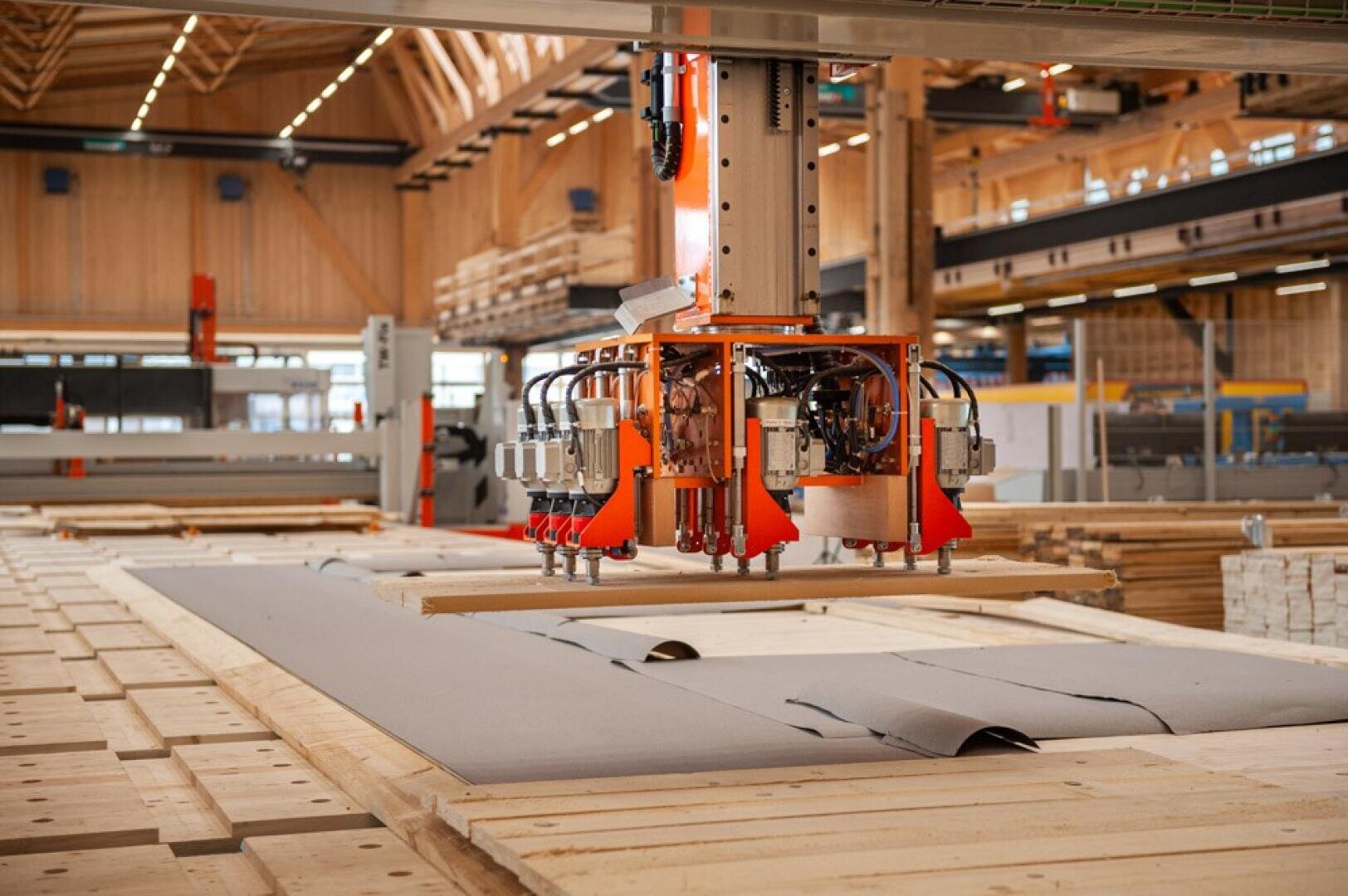 Un bras robotisé équipé de ventouses soulève et déplace de grands panneaux dans une installation de fabrication du bois spacieuse et moderne, avec des piles de bois et des lucarnes visibles.