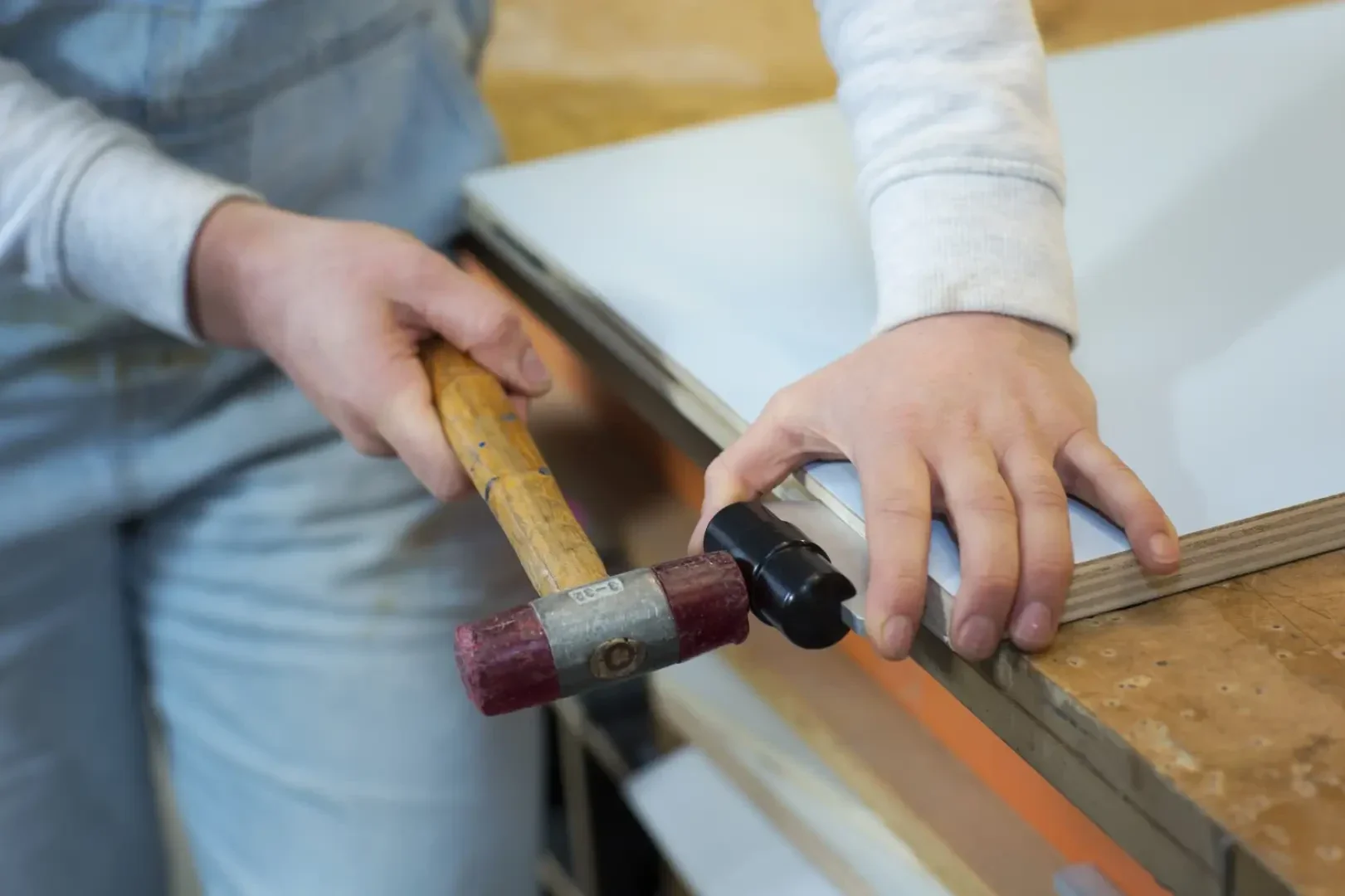 Eine Person verwendet einen Hammer, um eine schwarze Metallbefestigung an der Kante eines Holzbretts zu befestigen, wobei sie die Befestigung mit einer Hand festhält, während sie an einer Werkbank arbeitet.