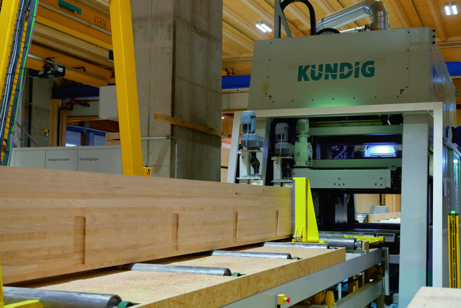 Eine große Kundig-Holzbearbeitungsmaschine bearbeitet einen langen Holzbalken in einer Industriehalle mit Sichtholzdecken und Betonpfeilern.