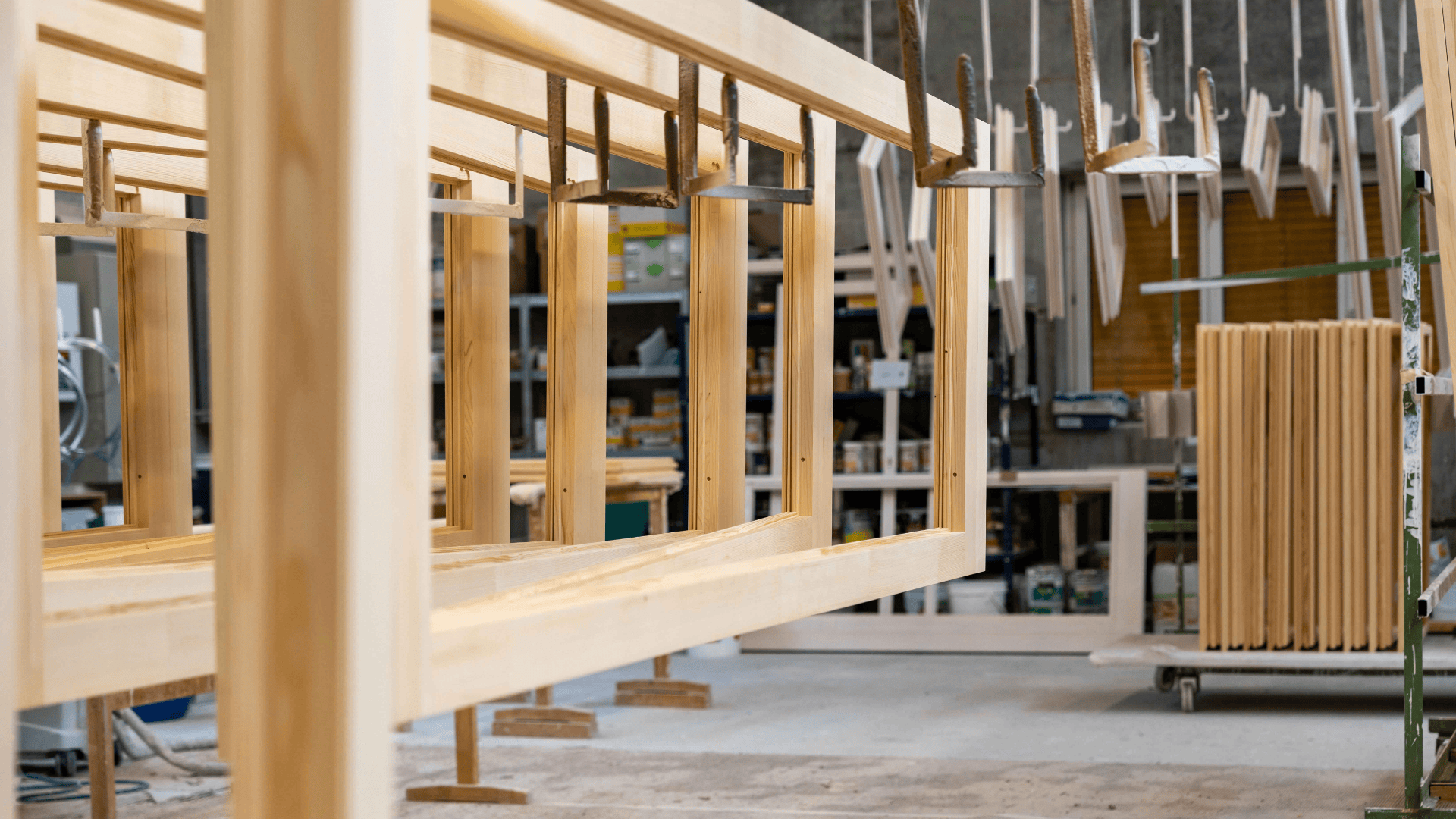 In einer Holzwerkstatt werden Holzrahmen zusammengespannt und aufgehängt, wobei im Hintergrund Werkzeuge, Regale und weitere Holzteile zu sehen sind. Die Szene deutet auf eine aktive Schreinerei oder Möbelherstellung hin.