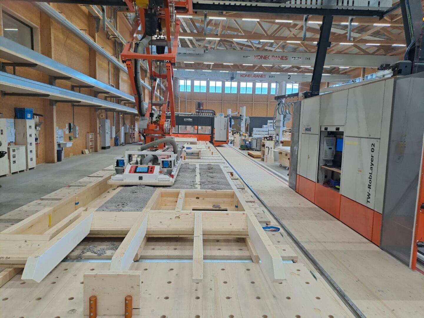 À l'intérieur d'une usine moderne, on peut voir des machines automatisées qui assemblent des panneaux muraux en bois. Des bras robotisés et des tapis roulants sont en action, et le bois et les matériaux de construction s'empilent dans l'espace de travail bien éclairé.