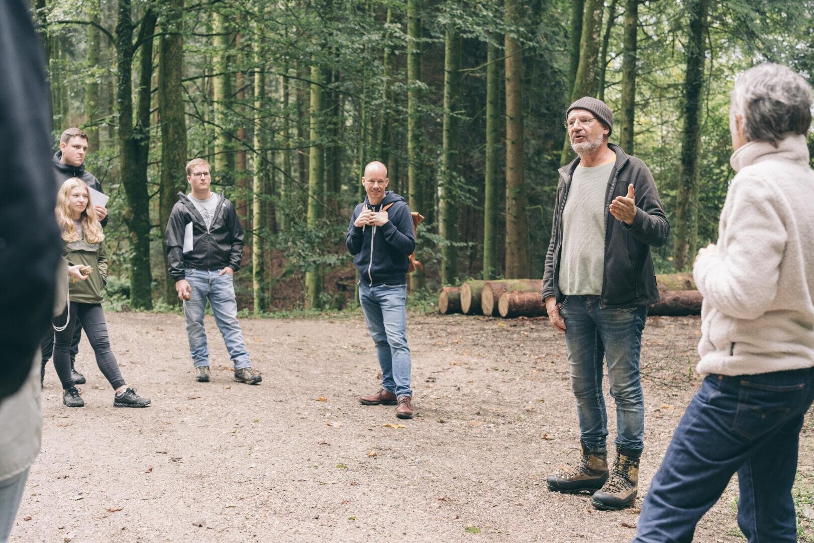 Eine Gruppe von Menschen steht auf einer Waldlichtung und hört einem Mann zu, der spricht. Die Gruppe wirkt engagiert, umgeben von hohen Bäumen und Baumstämmen, und alle sind in Freizeitkleidung gekleidet.