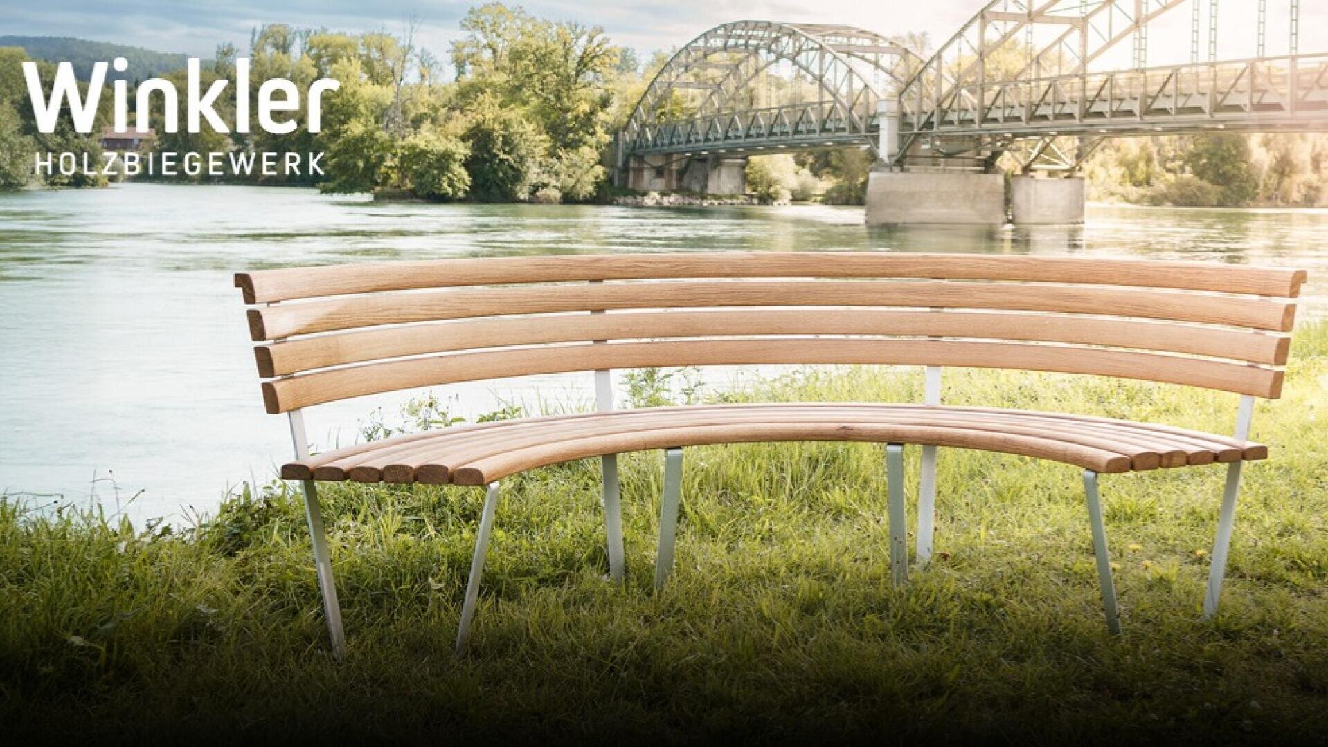 Eine geschwungene Holzbank mit Metallbeinen sitzt im Gras an einem Fluss, mit einer Stahlbogenbrücke im Hintergrund. Der Text Winkler HOLZBIEGEWERK erscheint in der oberen linken Ecke. Das Flussufer ist von Bäumen gesäumt.