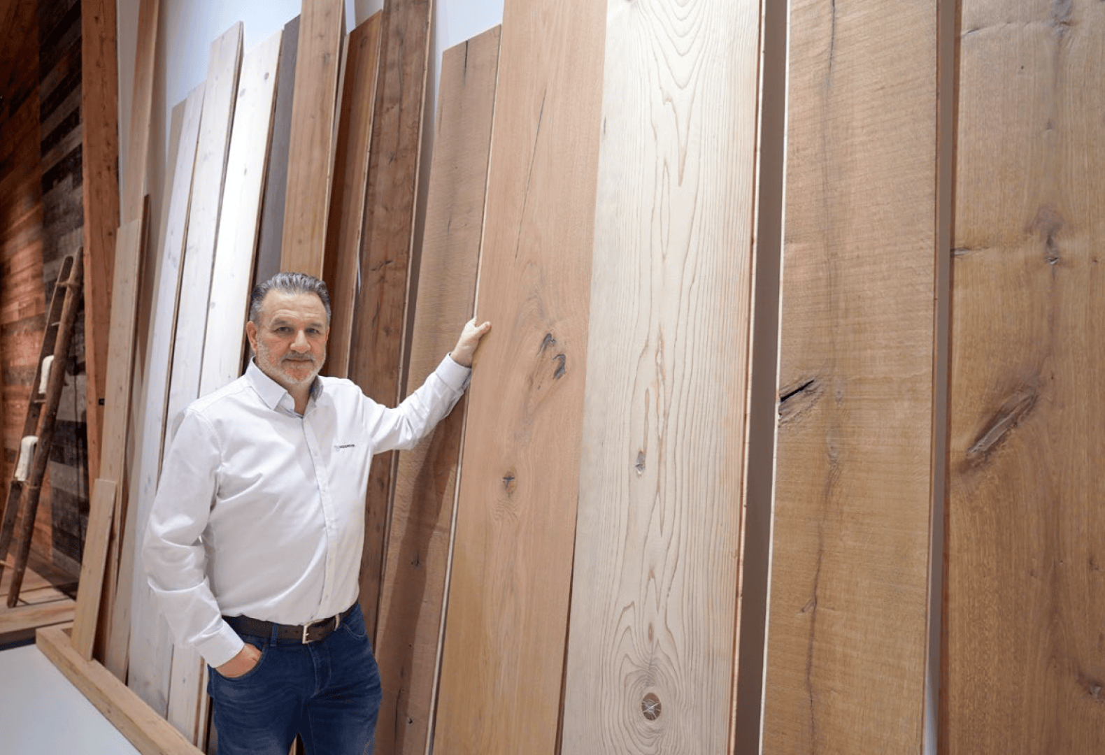 Un homme en chemise blanche se tient dans un intérieur et touche une grande planche de bois au milieu de plusieurs panneaux de bois verticaux fixés à un mur.
