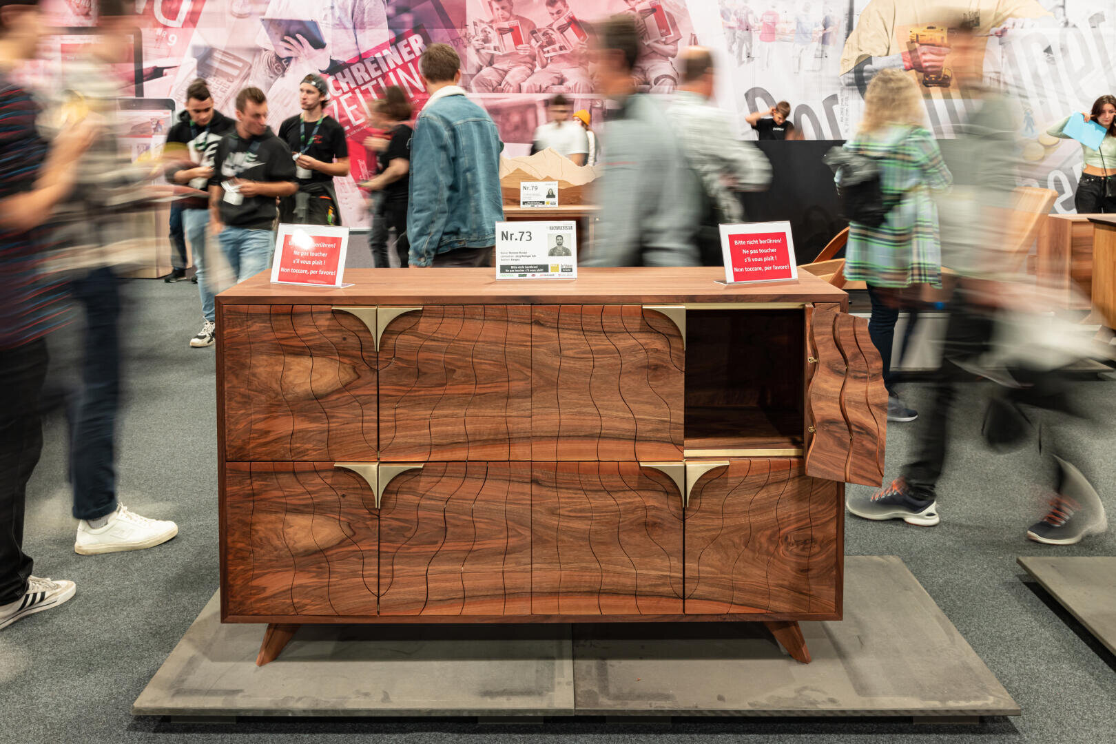 Ein hölzerner Schrank mit abstrakten geschnitzten Mustern und goldenen Akzenten wird in einer Ausstellung gezeigt. Menschen gehen vorbei und stehen herum, einige verschwommen in Bewegung, während Informationsschilder auf dem Schrank liegen.