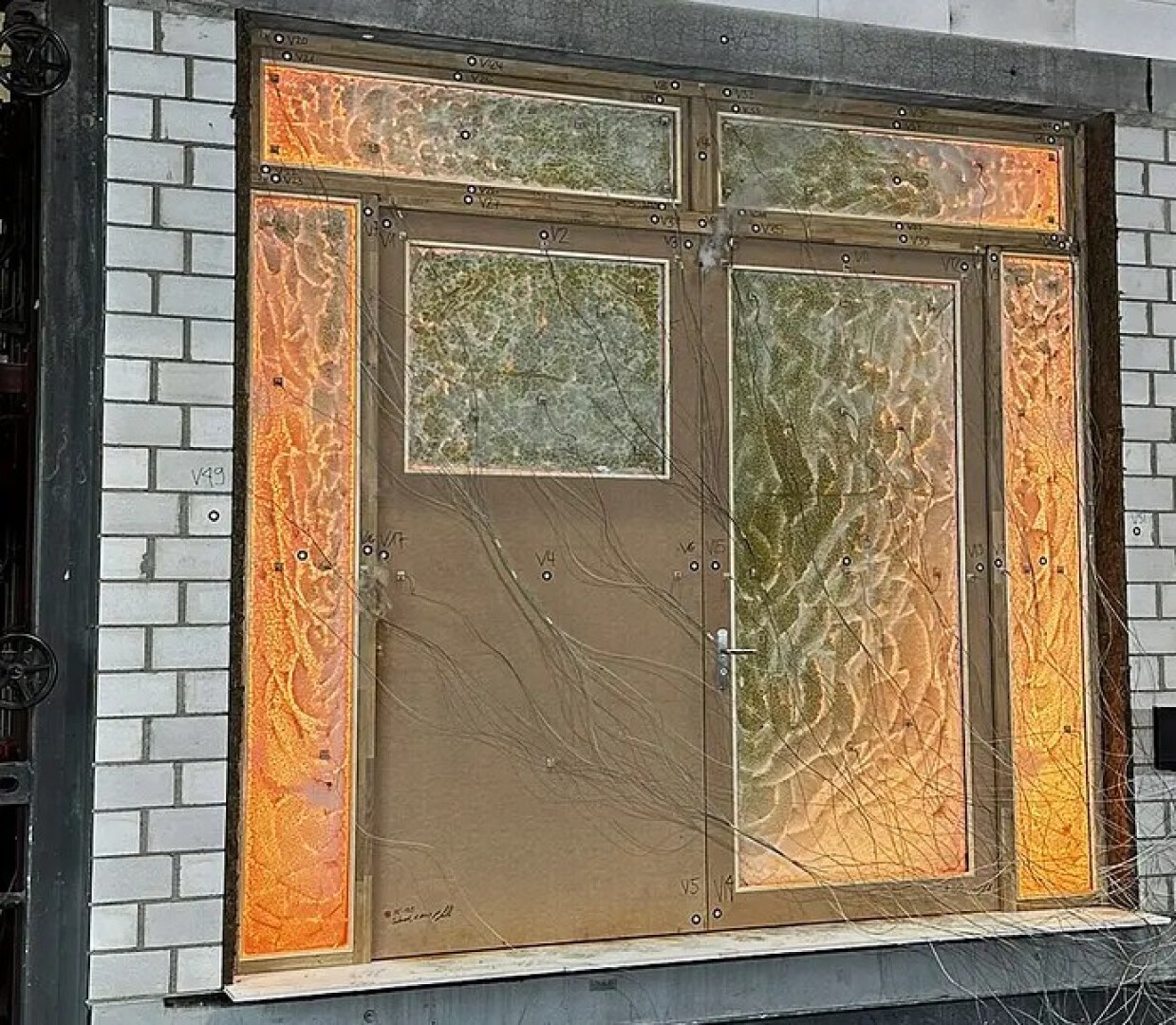 Eine Tür und Fenster, die mit undurchsichtigem, gemustertem Glas oder Folie bedeckt sind, leuchten von innen in warmem orangefarbenem Licht, eingebettet in eine weiße Ziegelwand. Vor dem Glas sind dünne Äste sichtbar.