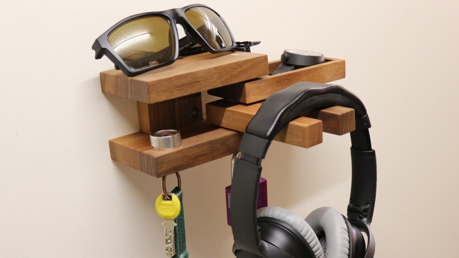 Un organisateur mural en bois contient des lunettes de soleil, une montre intelligente, une bague, des clés, un objet violet et une paire d'écouteurs. Les objets sont soigneusement exposés contre un mur clair et uni.