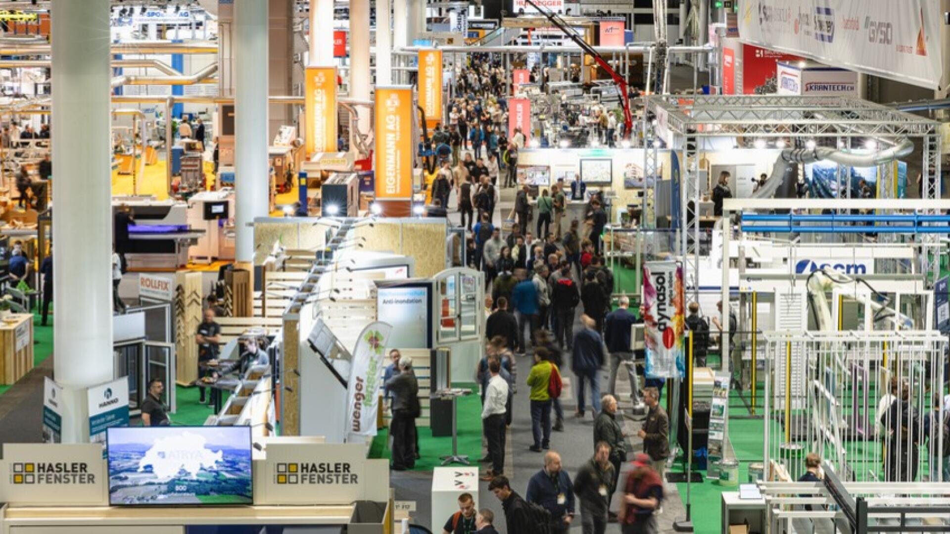 Un hall d'exposition de salon professionnel très fréquenté, rempli de stands, d'étalages et de nombreuses personnes qui se promènent et interagissent dans le lieu. De grandes enseignes et des logos d'entreprises sont visibles au-dessus des stands.