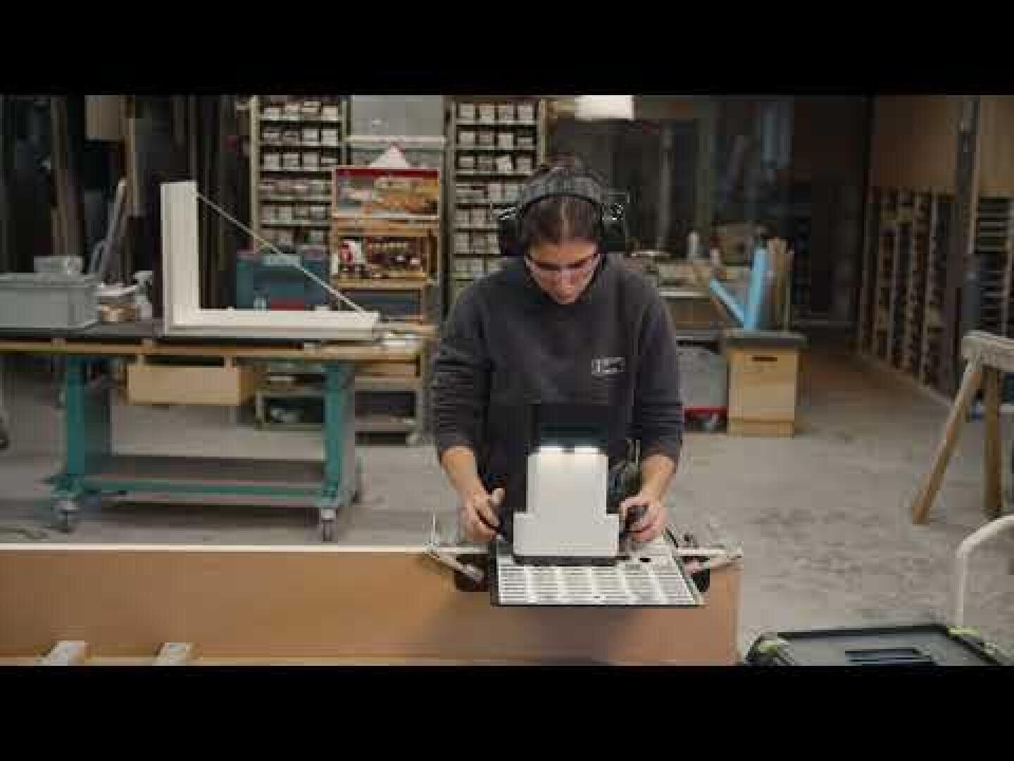 Une personne portant des lunettes de protection et des protections auditives utilise une machine dans un atelier de menuiserie, entourée d'étagères contenant des outils et des matériaux.