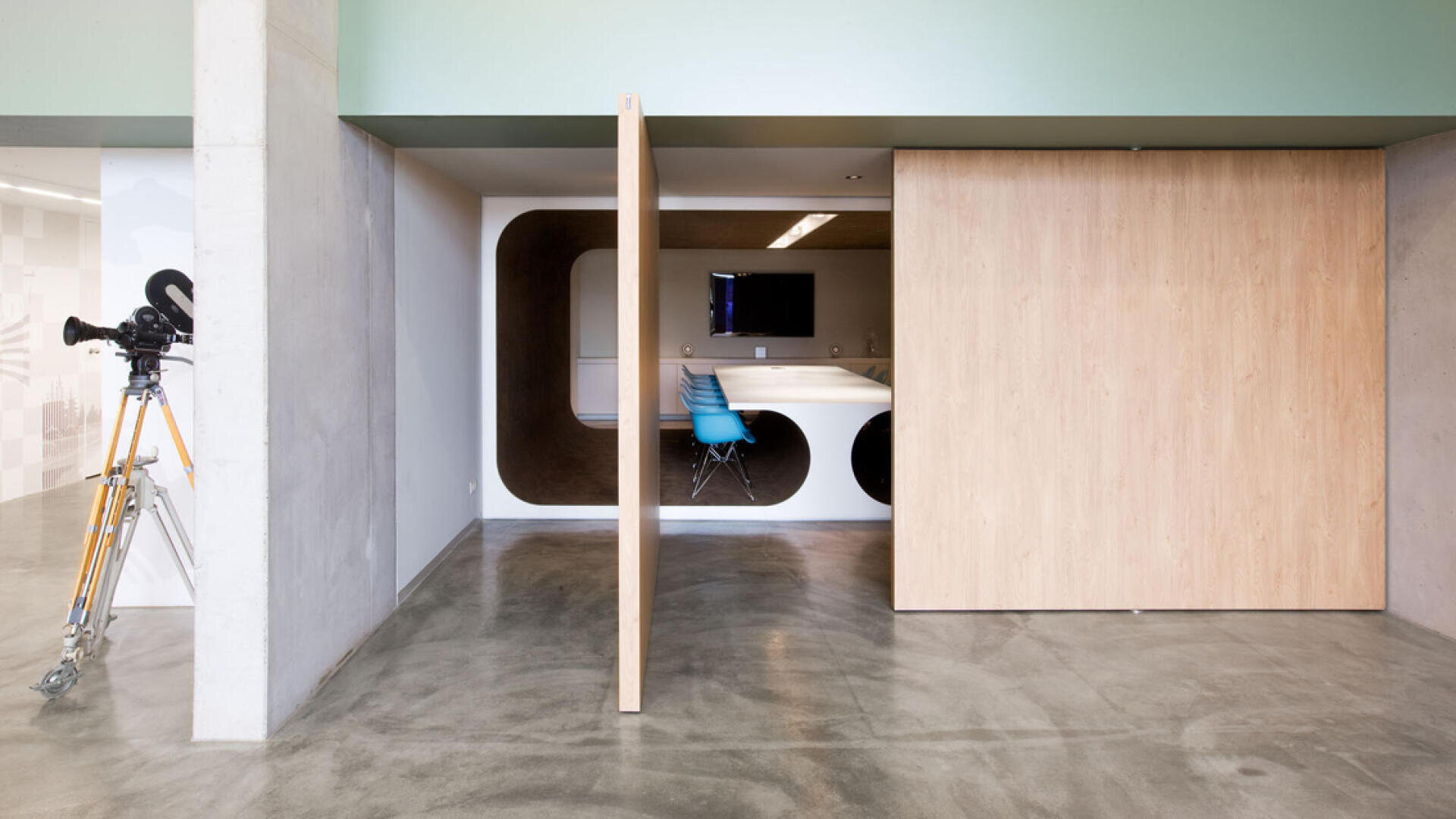 Moderne Büroräume mit polierten Betonböden, einer Schiebetür aus hellem Holz, die teilweise geöffnet ist und einen Besprechungsraum mit einem weißen Tisch, blauen Stühlen und einem an der Wand befestigten Bildschirm freigibt; draußen steht eine alte Filmkamera.