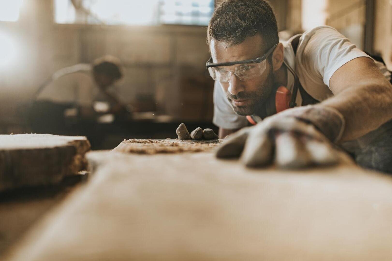 Ein Mann mit Schutzbrille und Handschuhen untersucht eine Holzoberfläche in einer Werkstatt, während im Hintergrund eine andere Person arbeitet und das Sonnenlicht einfällt.
