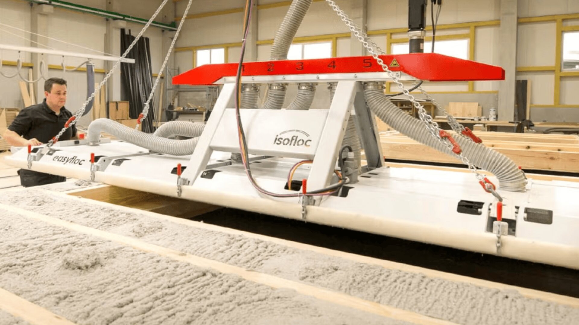 Ein Arbeiter bedient eine große Maschine mit der Bezeichnung isofloc, die in einer Werkstatt loses Dämmmaterial zwischen Holzbalken verteilt. Die Maschine ist an Kabeln aufgehängt und mit Schläuchen und einer roten Steuerleiste ausgestattet.