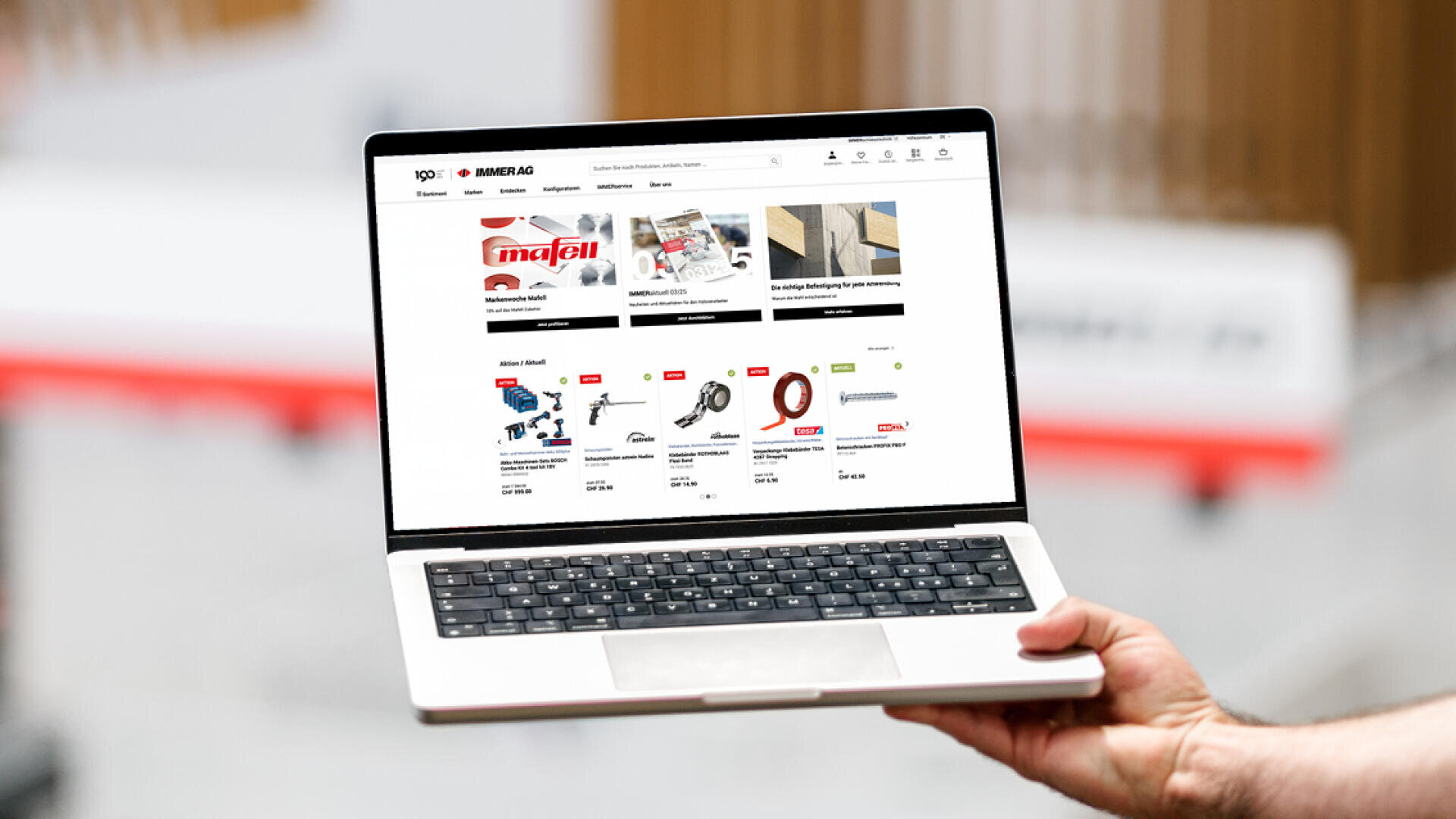 Eine Person hält einen Laptop in der Hand, auf dem eine Webseite eines Online-Shops angezeigt wird, auf der verschiedene Werkzeuge und Geräte zum Verkauf angeboten werden, darunter Sägen und Elektrowerkzeuge. Das Layout der Seite zeigt Produktbilder, Preise und Markennamen.