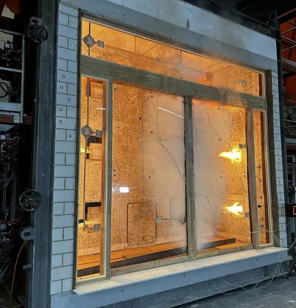 Ein großes Glasfenster wird im Labor auf Feuerbeständigkeit geprüft. Hinter dem Glas sind Flammen sichtbar, zur Überwachung sind Sensoren und Kabel am Fensterrahmen angebracht.
