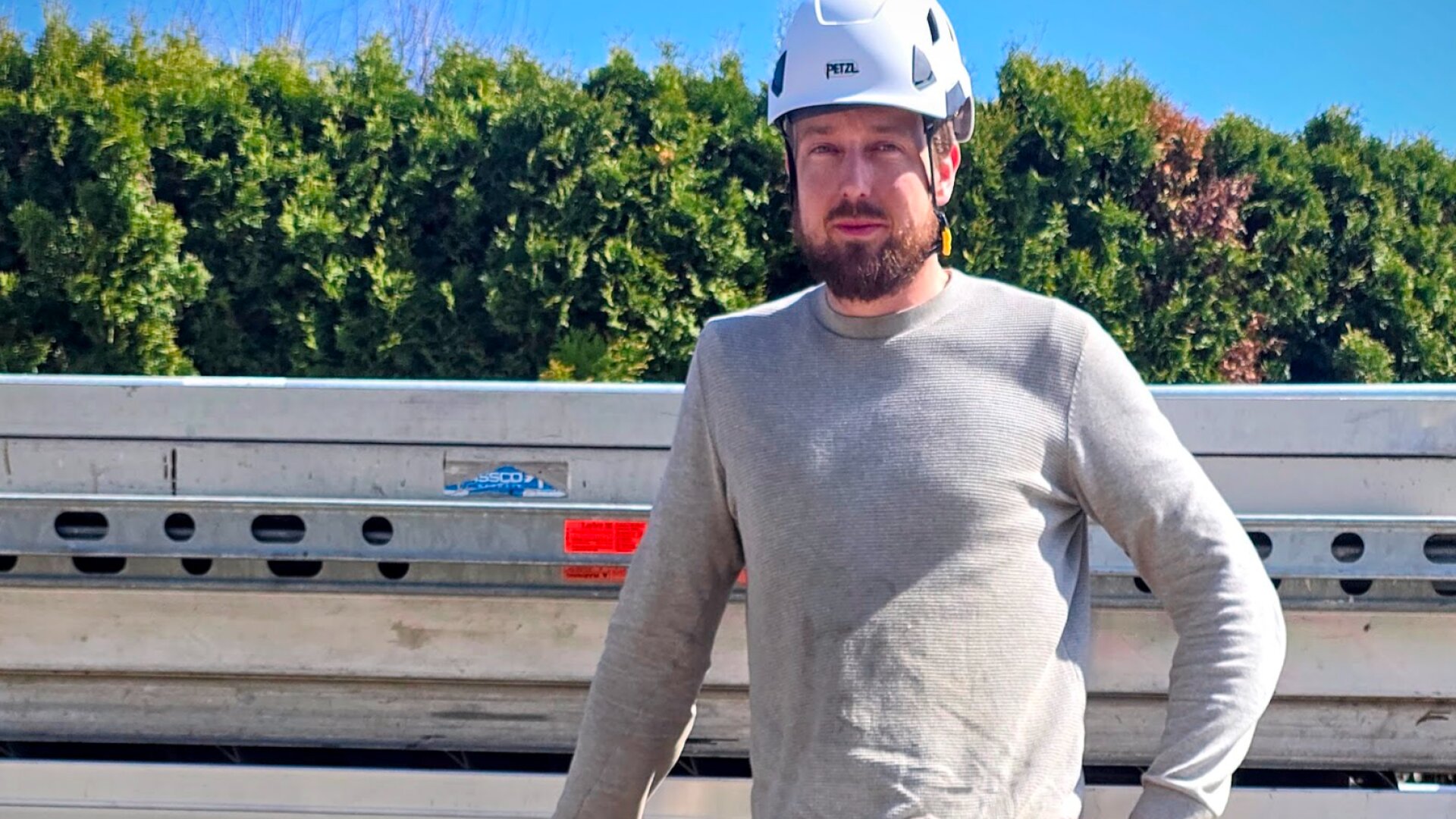 Ein Mann mit einem weißen Schutzhelm und einem hellgrauen langärmeligen Hemd steht im Freien vor einem gestapelten Metallgerüst, mit grünen Büschen und einem blauen Himmel im Hintergrund.