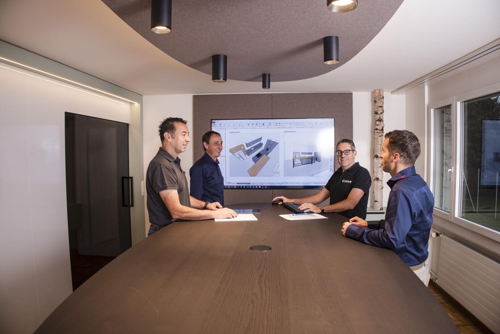 Quatre hommes se tiennent autour d'une table de conférence dans un bureau moderne et discutent de plans architecturaux qui s'affichent sur un grand écran derrière eux. Certains d'entre eux tiennent des papiers à la main et sourient. De grandes fenêtres assurent la lumière naturelle.