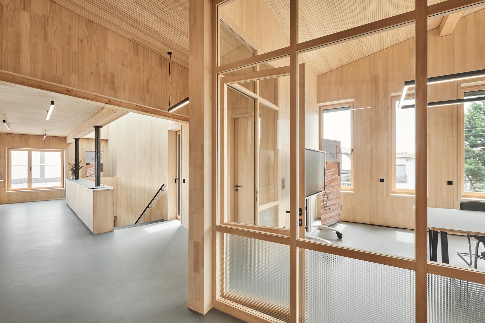 Moderne Büroeinrichtung mit hellen Holzwänden, Decken und Glastrennwänden. Große Fenster lassen natürliches Licht herein. Ein offener Arbeitsbereich und ein weißer Tresen mit schwarzen Armaturen sind zu sehen. Das Gesamtdesign ist minimalistisch und luftig.
