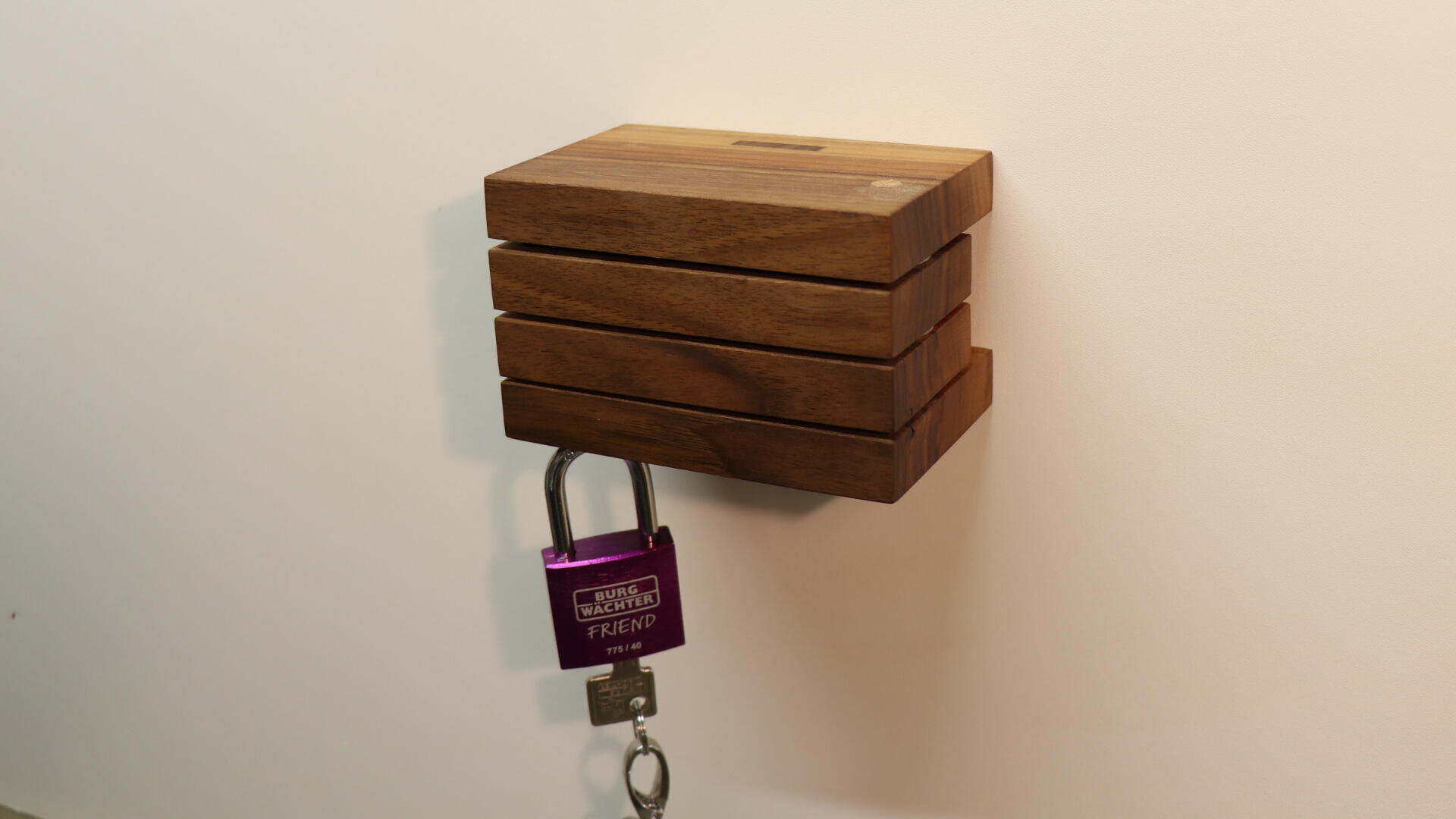 An einem hölzernen Wandkasten mit waagerechten Lamellen hängt ein violettes Vorhängeschloss an einem Metallhaken, zusammen mit einem Schlüssel an einem Schlüsselring. Der Kasten steht vor einer schlichten weißen Wand.
