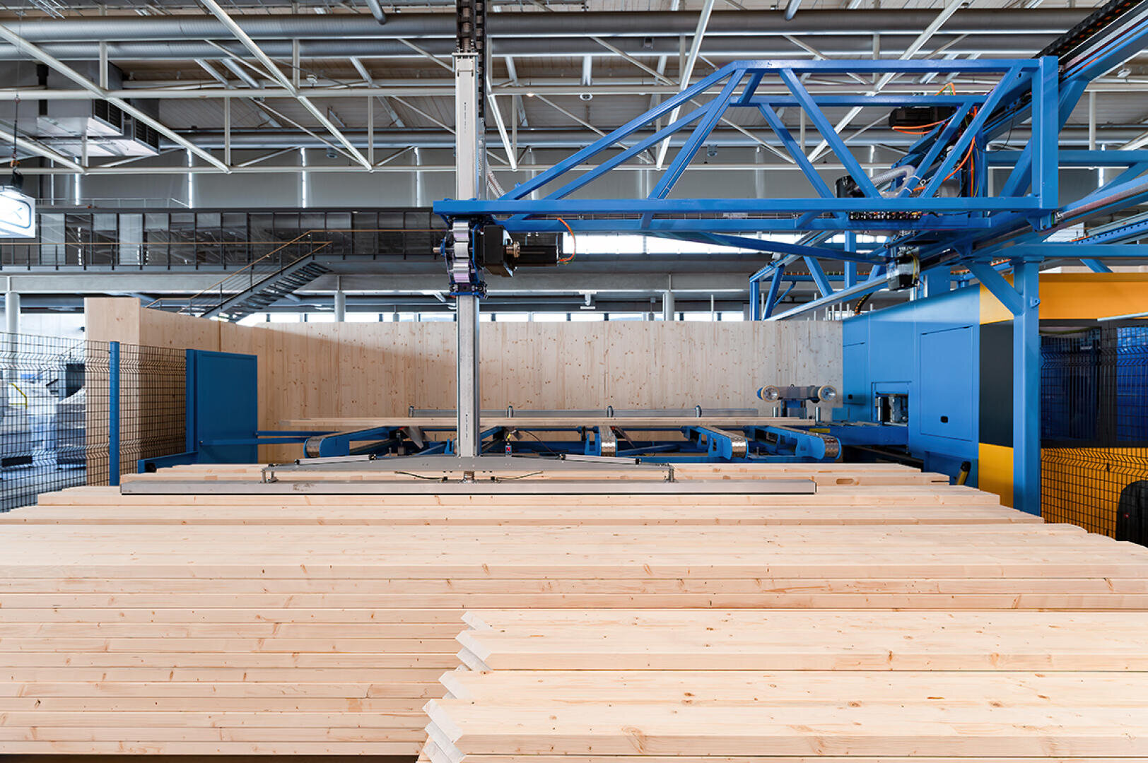 In einer modernen Industrieanlage sind Stapel heller Holzbretter fein säuberlich angeordnet. Im Hintergrund sind blaue Metallmaschinen und Oberlichter zu sehen.