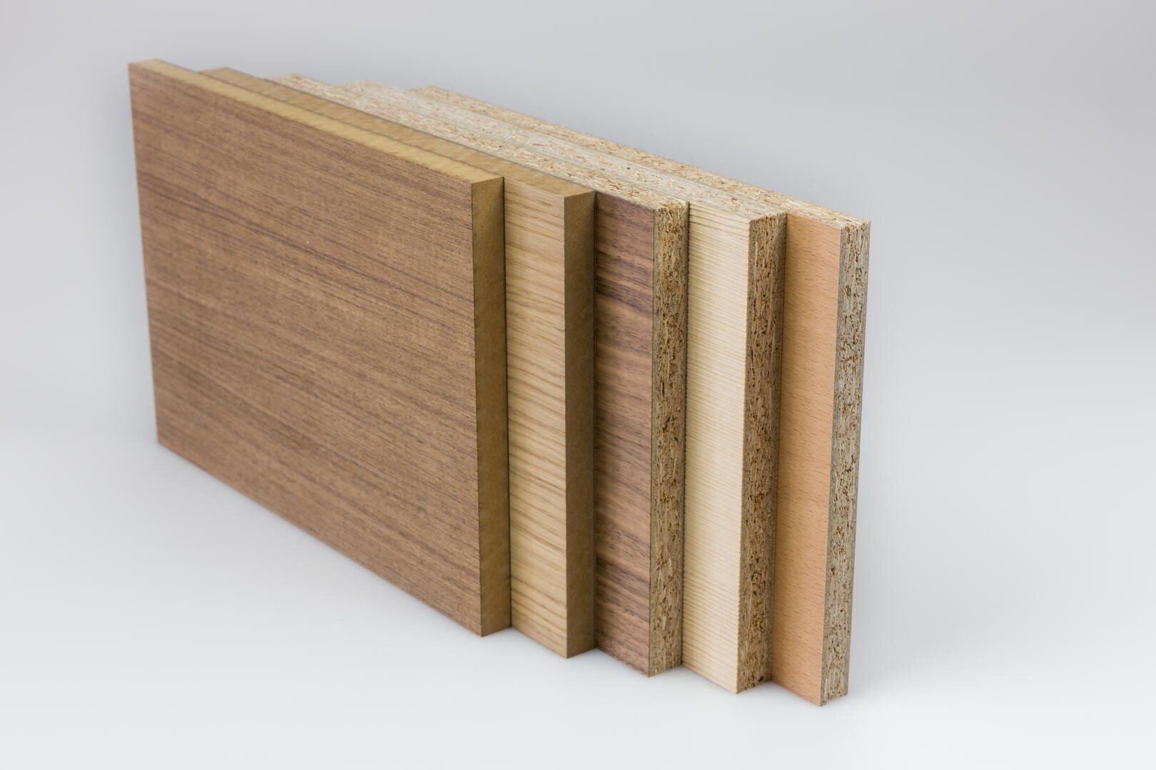 Cinq panneaux de bois rectangulaires d'épaisseur et de couleur différentes sont alignés verticalement sur un fond blanc et montrent leurs bords stratifiés et leurs différentes structures de surface.