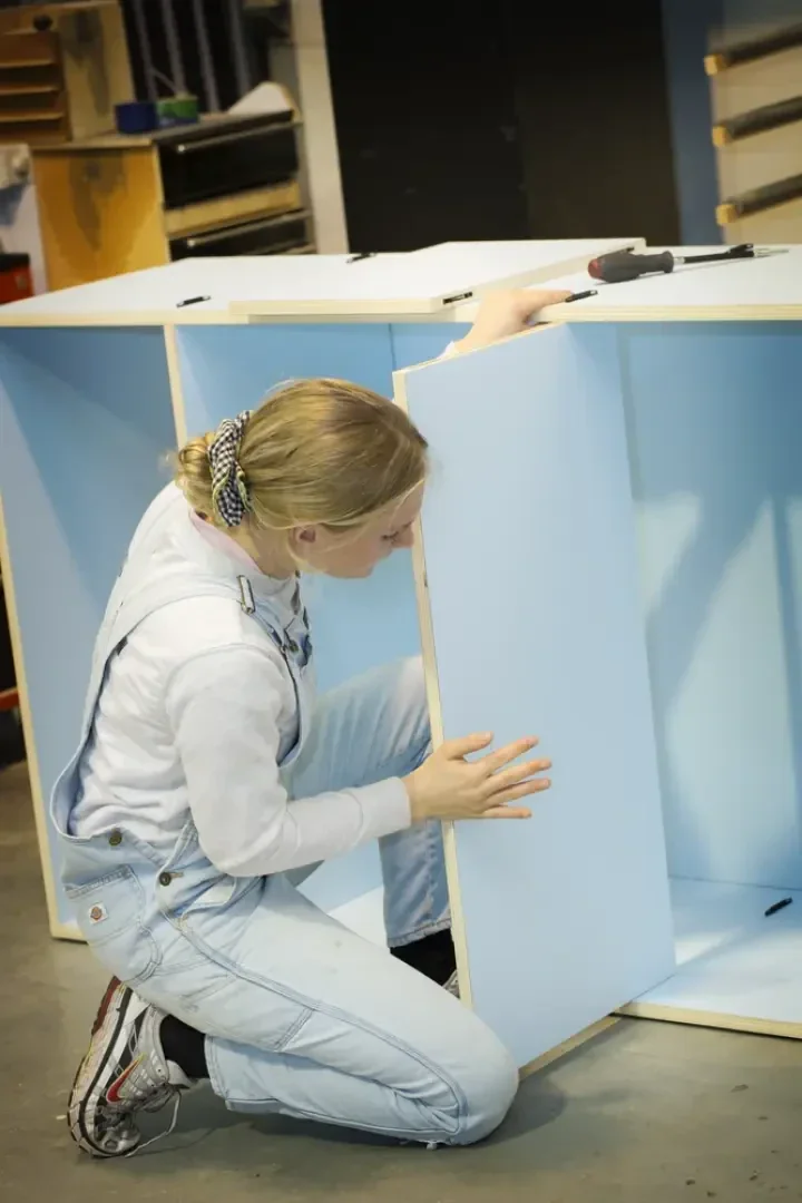Eine Person, die einen hellen Overall und einen Haargummi trägt, baut einen hellblauen Schrank zusammen, kniet auf dem Boden und justiert eine Platte. Im Hintergrund sind Werkzeuge und Regale zu sehen.