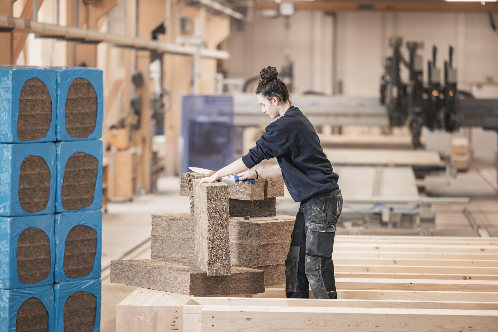 Une personne en tenue de travail empile des panneaux d'isolation bruns sur des poutres en bois à l'intérieur d'un atelier spacieux et bien éclairé. Des blocs d'isolation bleus sont visibles sur la gauche.