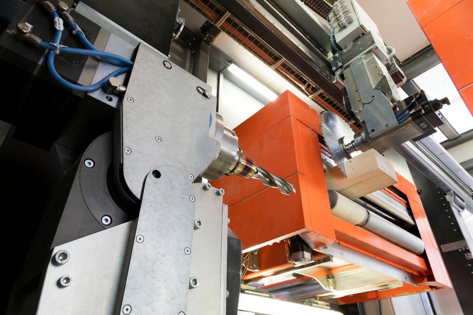Gros plan sur une machine industrielle équipée d'une grande perceuse à métaux et de composants orange, montrant un morceau de bois usiné avec précision dans un environnement de fabrication automatisé moderne.