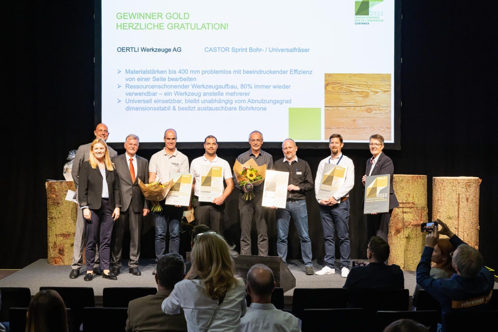 Eine Gruppe von Menschen steht auf der Bühne, hält Urkunden und Blumen in der Hand und posiert für ein Foto vor einer Präsentationsfolie, die einen Preisträger ankündigt, wobei mehrere Männer und Frauen in die Kamera lächeln.
