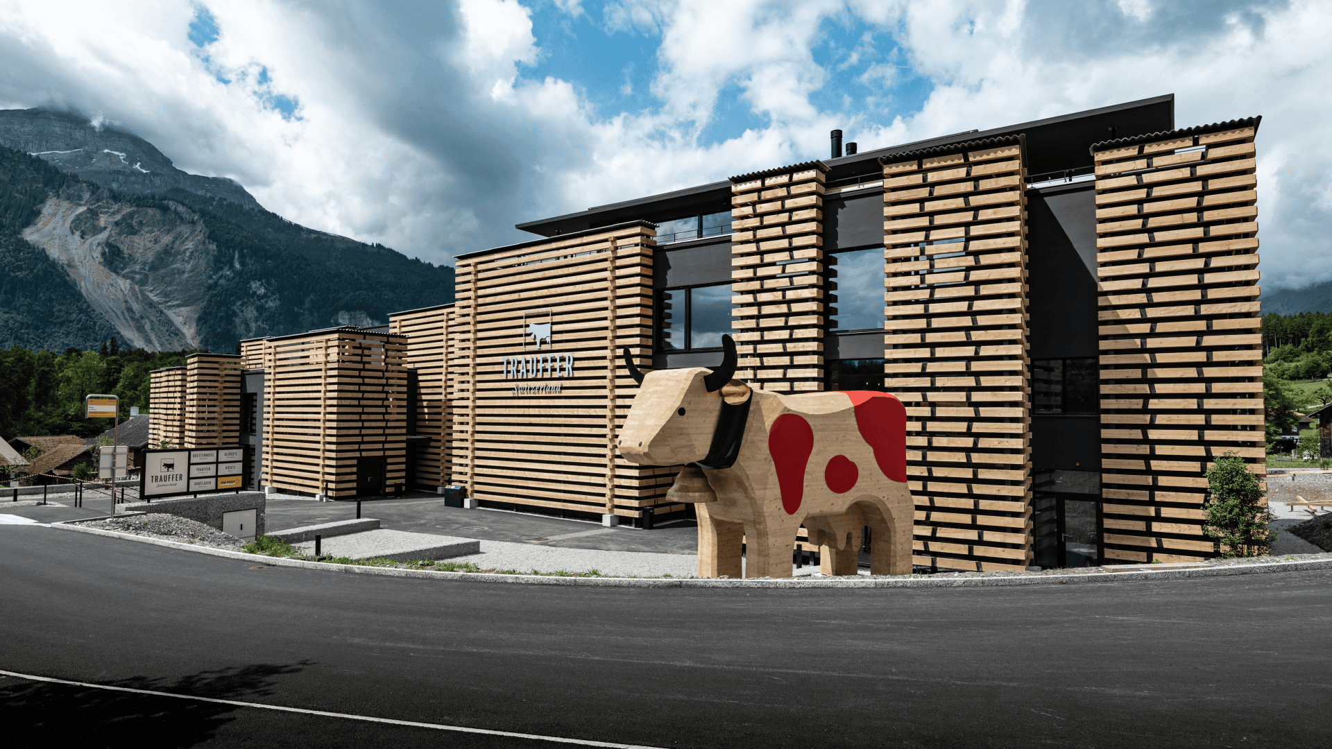 Ein modernes Gebäude mit horizontalen Holzlatten steht vor einer bergigen Kulisse. Davor steht eine große hölzerne Kuhskulptur mit aufgemalten roten Flecken. Der Himmel ist teilweise bewölkt.