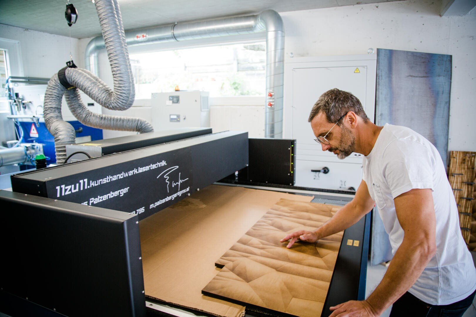Ein Mann mit Brille prüft eine strukturierte Holzplatte, die aus einer großen Industriemaschine in einer Werkstatt mit Metallrohren und Geräten im Hintergrund kommt.