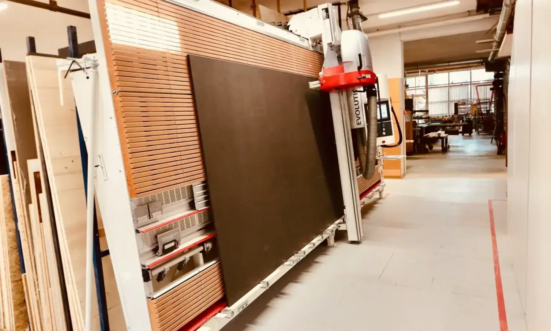 Une grande scie à panneaux verticale découpe un panneau rectangulaire sombre dans un hall industriel bien éclairé, avec des panneaux de bois et des équipements en arrière-plan.