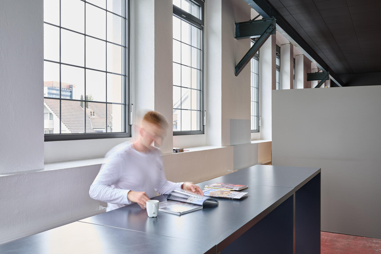 Eine Person in einem weißen Hemd sitzt an einem modernen Schreibtisch in der Nähe großer Fenster und blättert in Zeitschriften. Auf dem Schreibtisch steht ein Kaffeebecher. Die Umgebung ist hell, minimalistisch und industriell mit weißen Wänden und freiliegenden Stahlträgern.