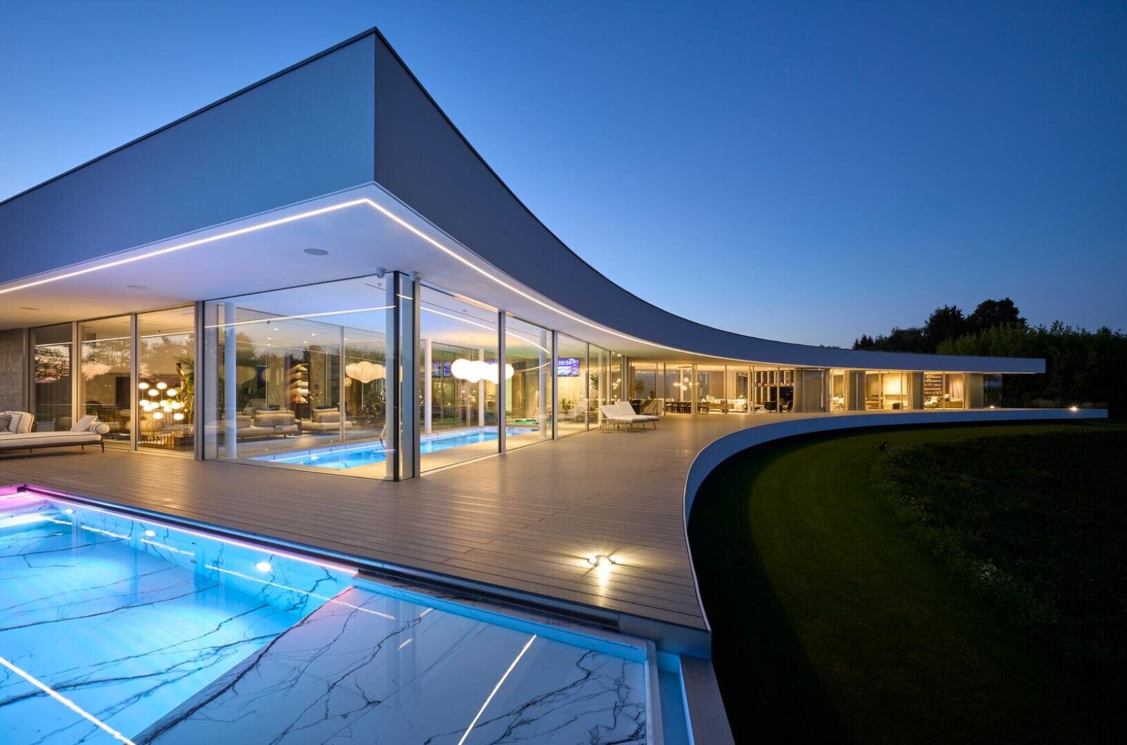 Modernes Luxushaus in der Abenddämmerung mit großen Glasfenstern, geschwungener Architektur und einem beleuchteten Außenpool mit Marmorakzenten. Innenleuchten beleuchten die eleganten Möbel, und ein gepflegter Rasen grenzt an die Terrasse.