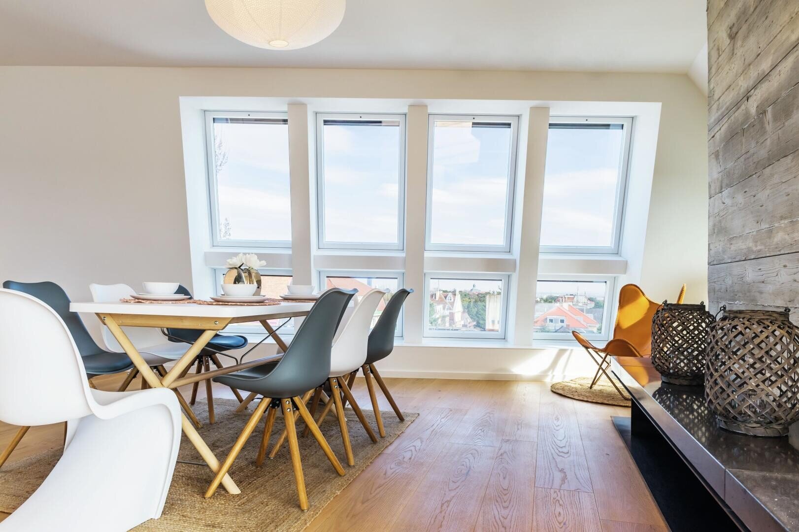 Modernes Esszimmer mit Holztisch, weißen und dunklen Stühlen, großen Fenstern, die natürliches Licht hereinlassen, gemütlicher Ecke mit gelbem Stuhl und dekorativen Laternen auf einem erhöhten Sims.