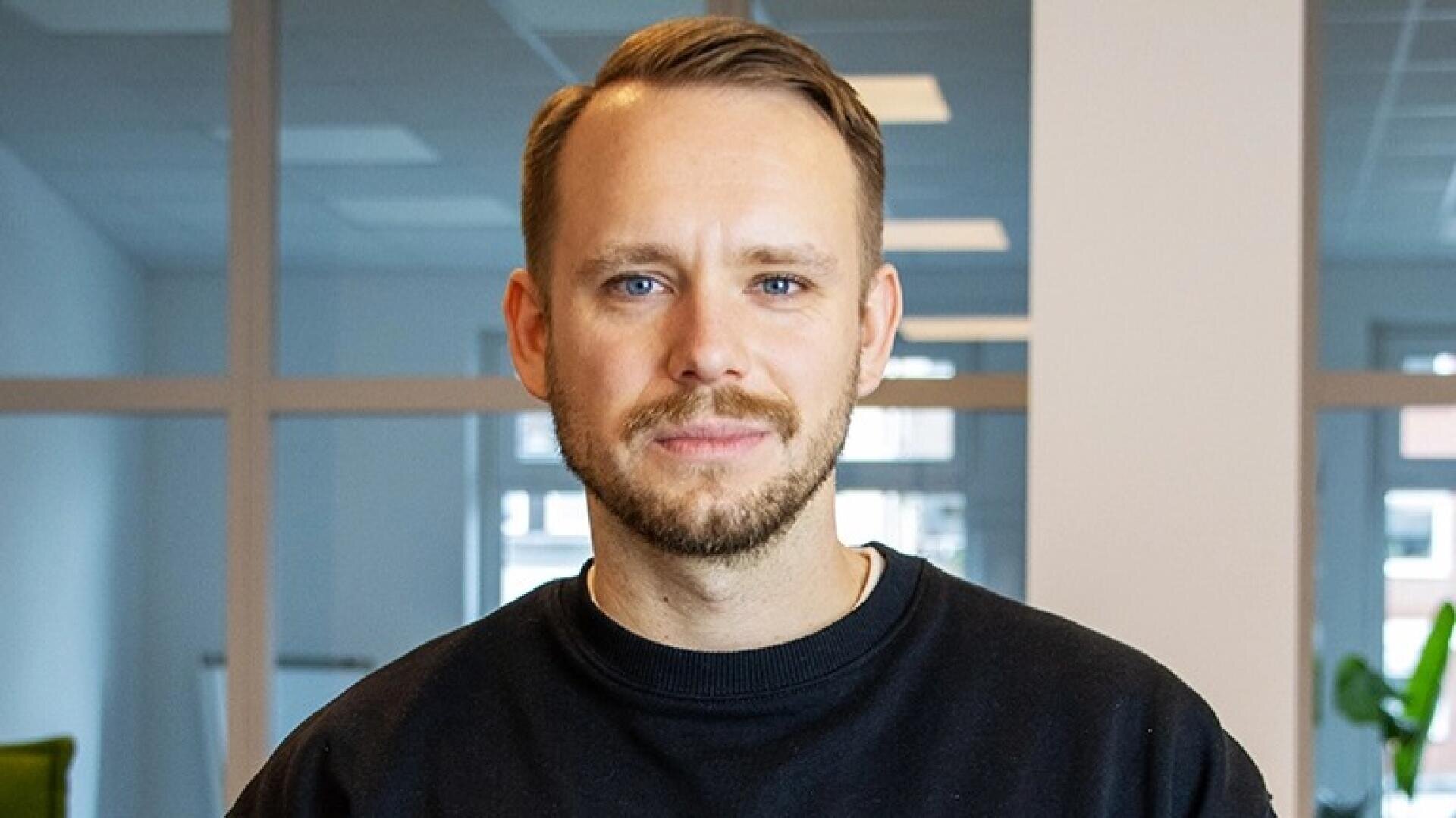 Ein Mann mit kurzen hellbraunen Haaren und gestutztem Bart, der ein schwarzes Sweatshirt trägt, steht in einem Innenraum vor einem Glasfenster und blickt mit neutralem Blick direkt in die Kamera.