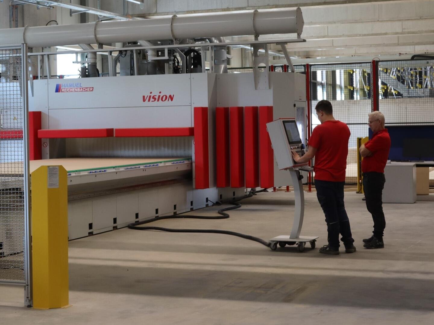 In einer geräumigen, modernen Fabrik bedienen zwei Männer in roten Hemden eine große Industriemaschine namens VISION. Einer von ihnen bedient ein Computerterminal, während der andere in der Nähe steht und beobachtet.