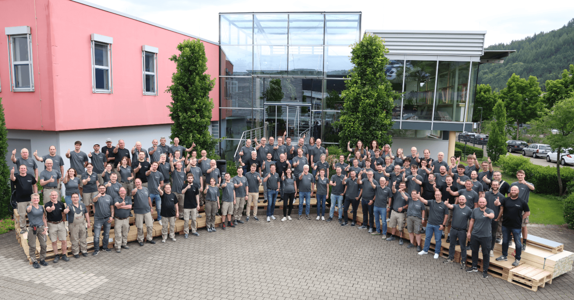 Eine große Gruppe von Menschen in passenden grauen Hemden und khakifarbenen Shorts oder Hosen posiert gemeinsam vor einem modernen Gebäude auf einer gepflasterten Fläche, die von Grünflächen und Bäumen umgeben ist.