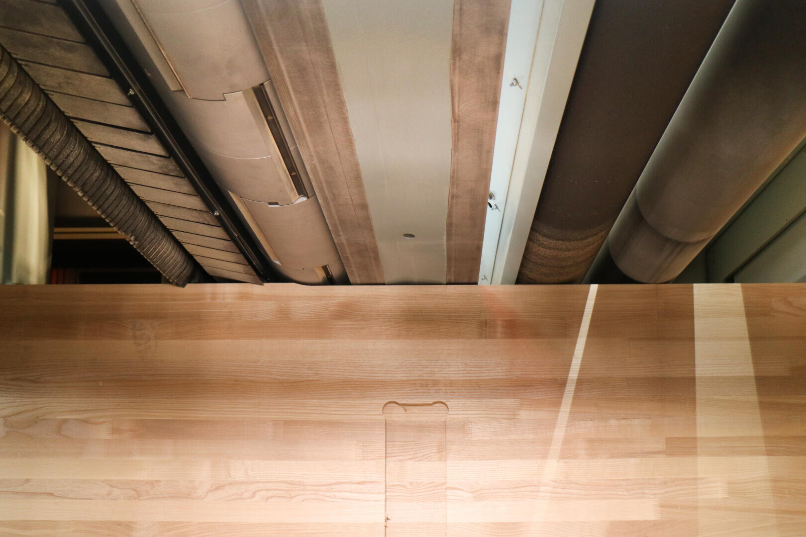 Ansicht einer glatten Holztischplatte mit hellen und dunklen Maserungen, von oben gesehen; im Hintergrund nahe der Decke verlaufen parallel dazu Industrierohre und Metallstrukturen.