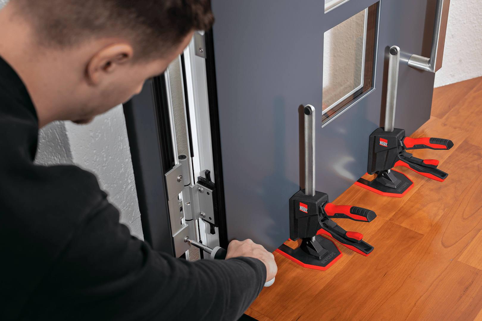 Une personne installe une porte à l'aide de deux outils de serrage noirs et rouges pour la fixer sur une surface en bois, tout en ajustant l'orientation de la porte à l'aide d'un tournevis.