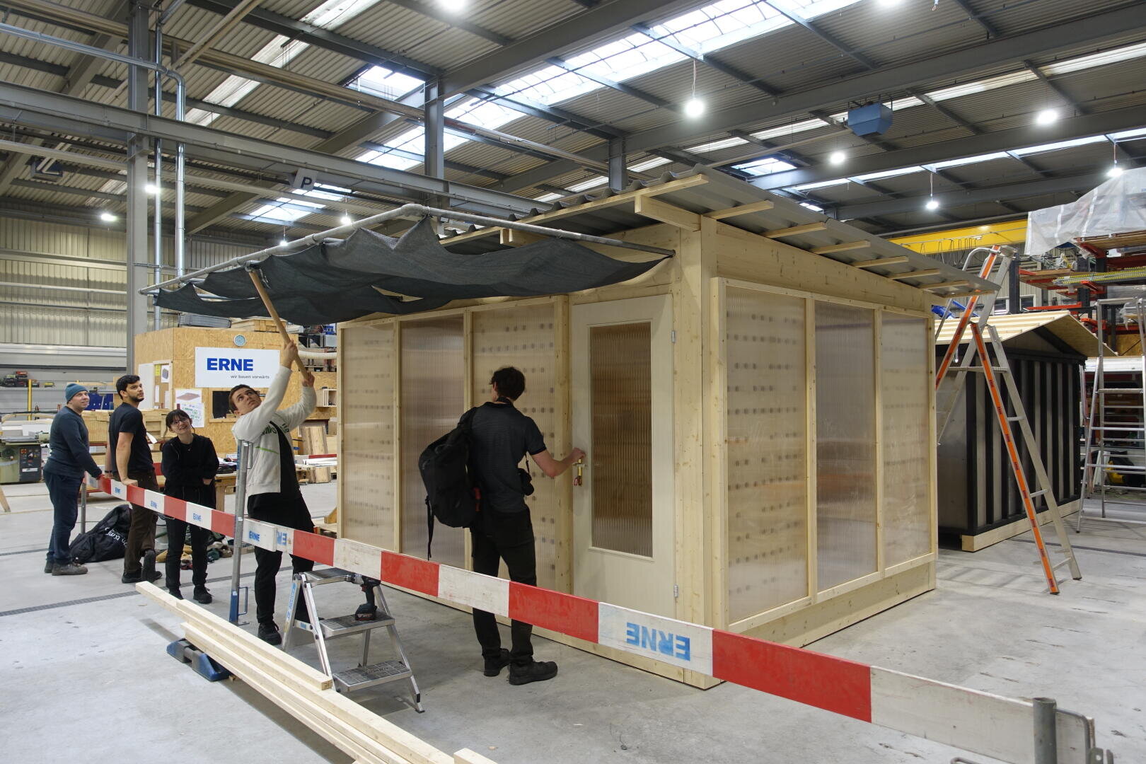 Eine Gruppe von Menschen baut in einem Haus eine kleine Holzkonstruktion zusammen. Einige arbeiten mit Werkzeugen, während andere zusehen. Eine schwarze Plane ist teilweise auf dem Dach angebracht, und rot-weißes Klebeband sperrt den Arbeitsbereich ab. Der Schauplatz ist eine große Industriehalle.