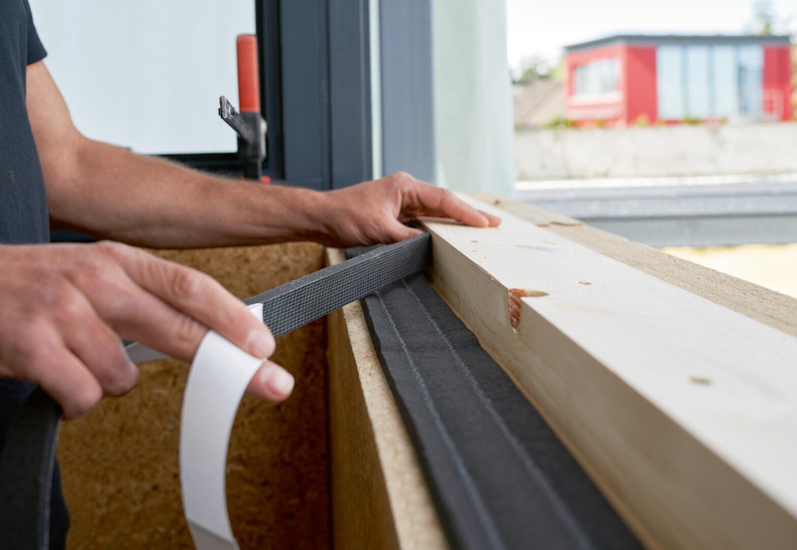 Une personne colle du ruban adhésif sur une poutre en bois près d'une fenêtre, peut-être pour préparer l'isolation ou l'étanchéité lors de travaux de construction ou de rénovation.