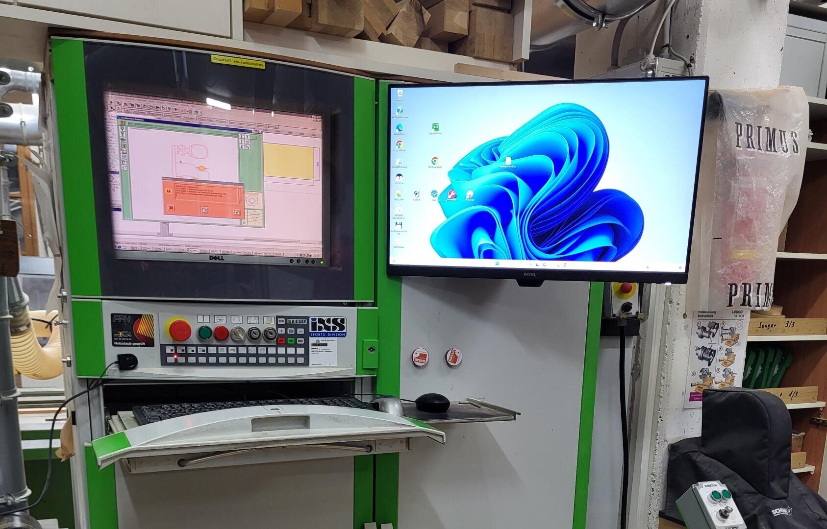 Eine CNC-Maschine für die Holzbearbeitung mit einem Bedienfeld und zwei Bildschirmen: einer zeigt die CNC-Software an, der andere den Windows 11-Desktop. Die Maschine steht in einer Werkstatt, in der Werkzeuge und Materialien herumliegen.