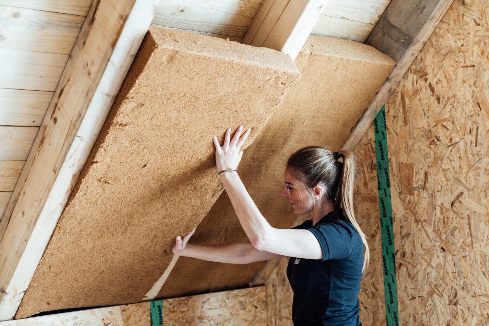 Eine Frau bringt eine große Platte aus Dämmmaterial zwischen den Holzbalken einer schrägen Dachgeschossdecke in einem im Bau befindlichen Haus an.