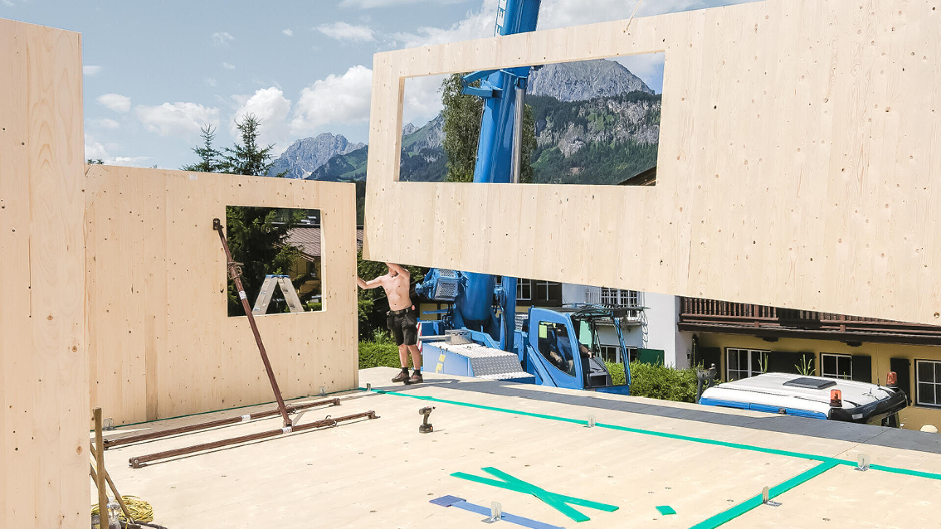 Ein Bauarbeiter steht auf einer Holzbaustelle, während ein Kran eine große vorgefertigte Wandplatte an ihren Platz hebt. Im Hintergrund sind Berglandschaften und Bäume bei teilweise bewölktem Himmel zu sehen.